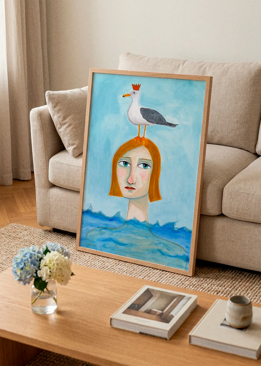 Crowned Seagull Woman by Sharyn Bursic displayed in a natural wood frame, leaning against a beige sofa in a warm, softly styled living room with a wooden coffee table.