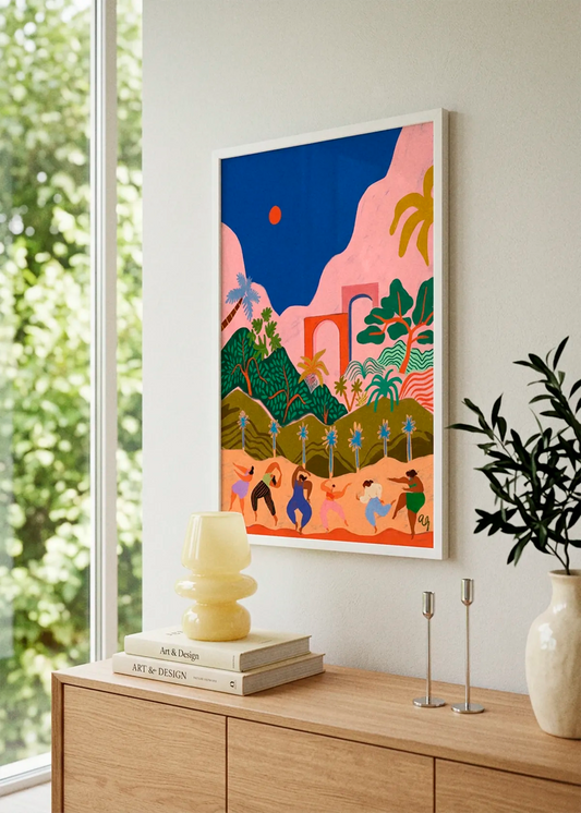 Dancing In Paradise by Arty Guava art print featuring dancing figures, lush green hills, palm trees, pink mountains and a deep blue sky, displayed in a white frame in a bright Scandinavian interior.