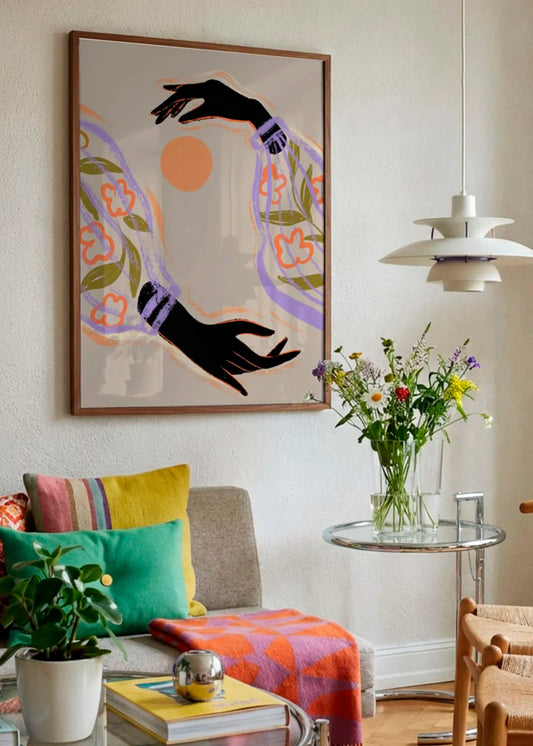 Indrammet farverig kunstplakat med hænder, blomster og sol, hængt i en lys stue med farverige tekstiler og blomster.