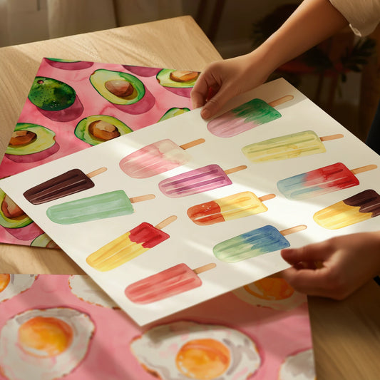 Hands holding a colorful popsicle art print, showing the matte high-quality paper texture, with Avocado and Fried posters lying on the table in a warm kitchen setting.