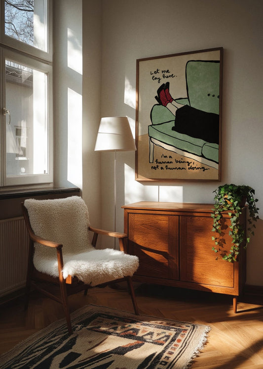 Let Me Lay Here poster in an oak frame hanging in a cozy Scandinavian living room corner, with warm natural sunlight, a vintage wooden cabinet, a soft sheepskin chair, and indoor greenery.