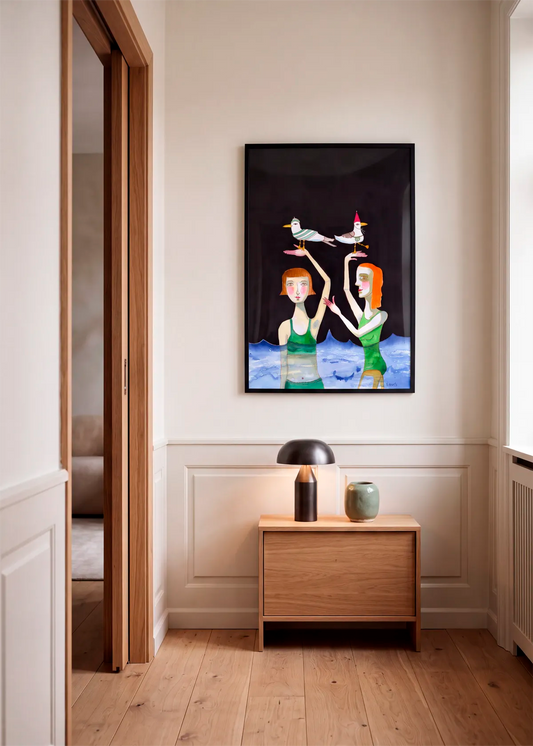 Seagulls In Hats by Sharyn Bursic – framed art poster featuring two women in green swimsuits standing in blue water with seagulls wearing hats, styled in a warm neutral hallway.