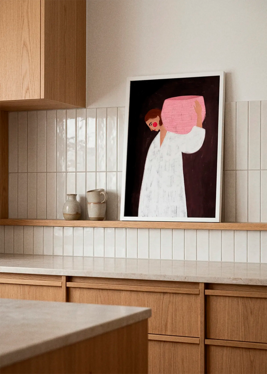 Serene Woman Carrying Basket by Bea Müller displayed in a white frame on a wooden kitchen shelf, styled with handmade ceramic vases and warm oak cabinetry in a calm, modern interior.