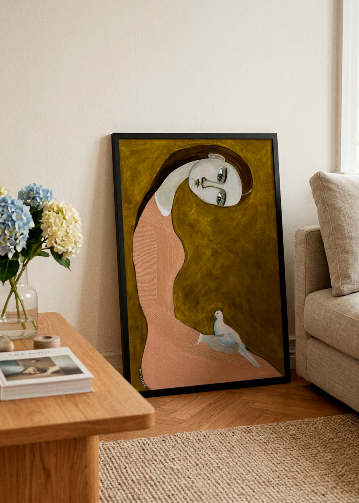Serene Woman and Dove by Sharyn Bursic displayed in a black frame, leaning against a soft cream wall beside a beige sofa and wooden coffee table in a warm, elegant living room.