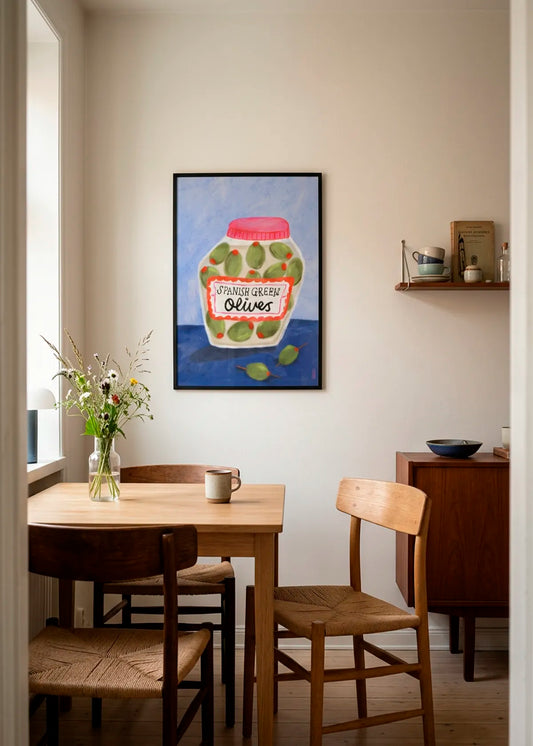 Spanish Green Olives Jar by Dirkie Nell – framed art poster with a hand-painted jar of green olives, pink lid and blue background, styled in a warm Scandinavian dining room with wooden furniture.