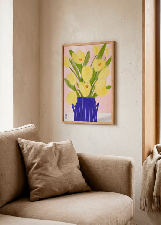 Spring Tulips Blue Vase by Dirkie Nell – framed floral poster with soft yellow tulips, green leaves and a cobalt blue vase on a pale pink background, styled above a neutral armchair.