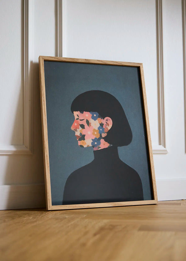 A Face I Once Knew portrait poster in an oak frame, styled against a white panelled wall with a floral female profile on a dark blue-grey background.
