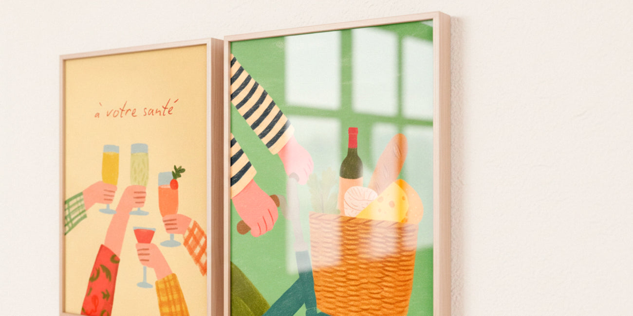 Two framed posters from the French Picnic collection hanging on a light wall, featuring illustrated hands toasting drinks and a cozy picnic basket scene with warm daylight reflections.