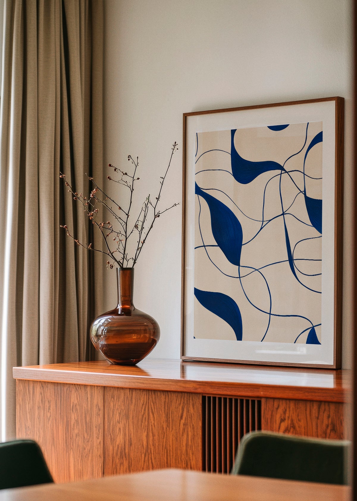 A framed poster titled "Art" featuring navy blue abstract curves on a beige background, styled on a wooden sideboard with a brown glass vase and branches, in a modern Nordic interior with beige curtains.