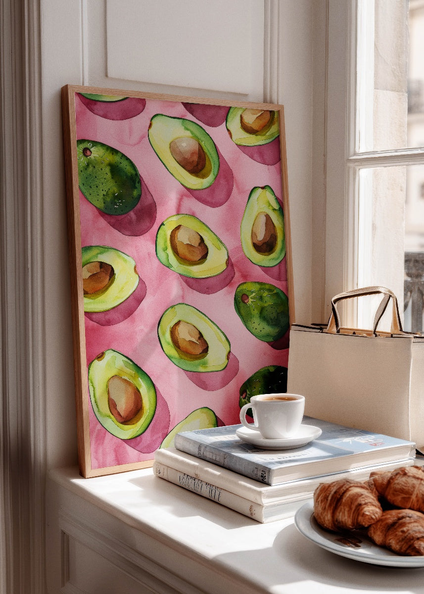 Framed avocado art print on a pink background, styled on a windowsill with books, coffee, and croissants – perfect for kitchen or breakfast nook wall decor.