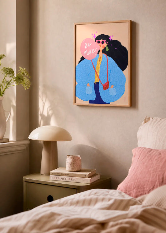 Be Nice Bubblegum Girl by Dirkie Nell – framed colourful art poster with a stylish woman in a blue jacket holding a pink “Be Nice” sign, styled in a warm bedroom interior.