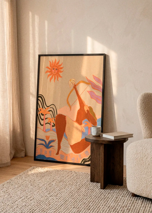 Bow And Arrow by Arty Guava art print featuring a woman drawing a bow beneath a radiant sun, surrounded by palm trees and flowing shapes in warm peach, orange and blue tones, displayed in a black frame in a sunlit Scandinavian interior.
