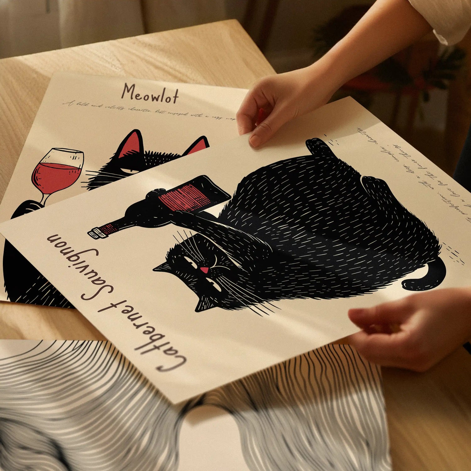 Hands lifting the Catbernet Sauvignon cat poster, highlighting the matte art paper and detailed illustration with warm indoor lighting.
