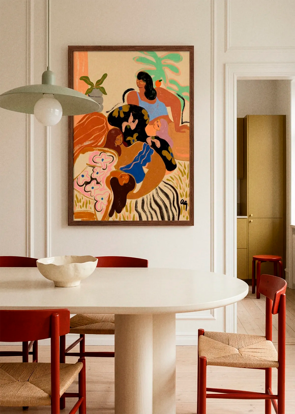Cozywozy by Arty Guava art print featuring three women relaxing together in a warm, colourful interior, displayed in a dark wood frame above a dining table with red chairs.