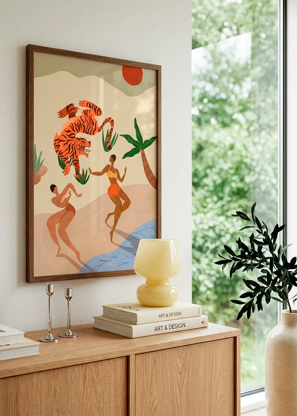 Dancing Tiger by Arty Guava art print featuring two women dancing beneath a leaping orange tiger, with tropical plants, a red sun and soft blue water, displayed in a dark wood frame in a bright Scandinavian interior.