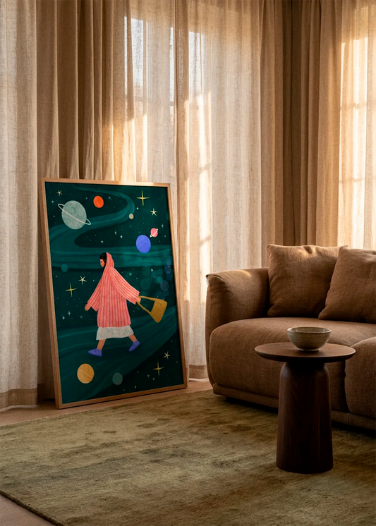 Dreamy Space Walk by Bea Müller displayed in a natural wood frame, leaning against soft beige curtains beside a brown sofa in a warm, sunlit living room.