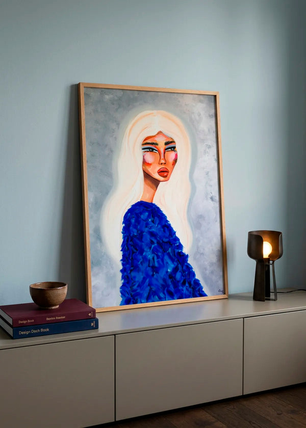 Elegant Blue Fashion by Amy Straley – framed portrait art poster featuring a woman with long white hair and a vivid textured blue top, styled on a modern sideboard against a soft blue wall.