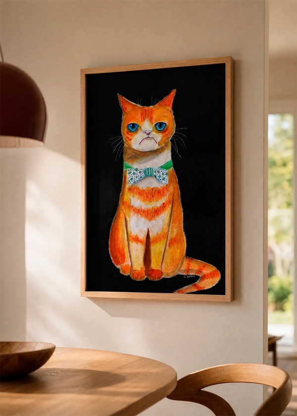 Grumpy Cat Bow Tie by Sharyn Bursic displayed in a natural wood frame on a soft beige wall, styled above a wooden dining table in a warm, sunlit interior.