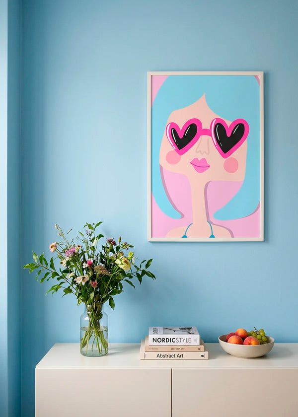 Heart Glasses by Athene Fritsch – framed illustrative portrait poster with blue hair and hot pink heart-shaped sunglasses, styled on a bright blue wall above a white cabinet.