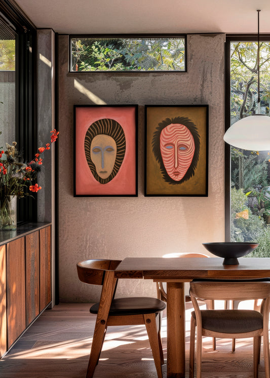 Framed posters Junglemen 1 and Junglemen 2 hanging side by side in a modern dining room with natural light, wooden furniture, and large windows. The left poster features a coral red background and striped mask motif, while the right one shows a mask with wavy lines on a golden brown background.