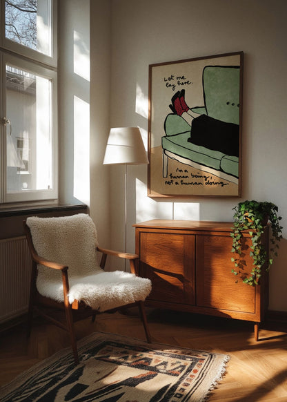 Let Me Lay Here poster in an oak frame hanging in a cozy Scandinavian living room corner, with warm natural sunlight, a vintage wooden cabinet, a soft sheepskin chair, and indoor greenery.