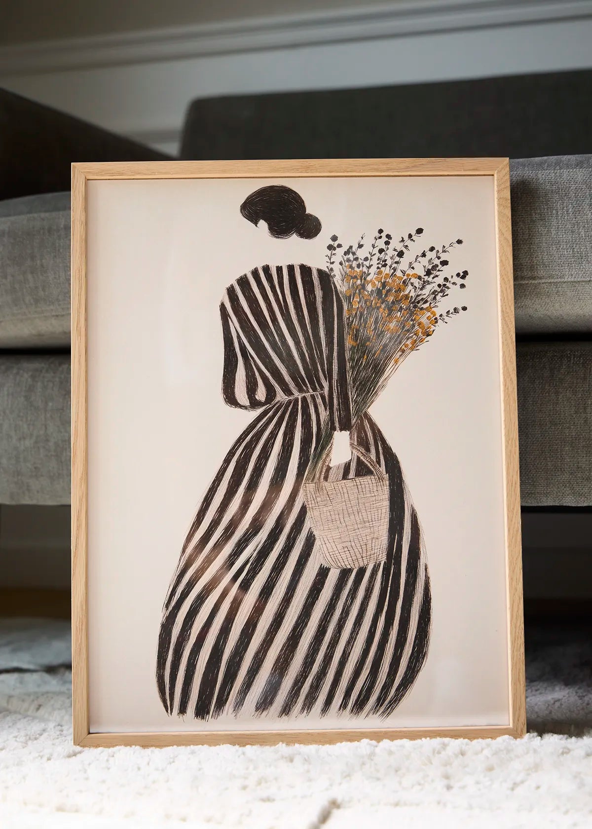 MariGold floral poster in an oak frame styled beside a grey sofa, featuring a woman with wildflowers in black, beige and warm yellow tones.