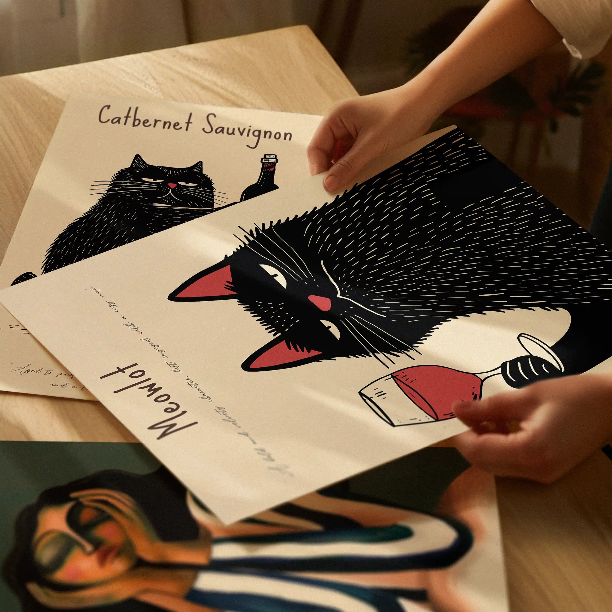 Hands holding the Meowlot wine-cat poster, showing the matte paper texture and rich print quality, with the Catbernet print lying underneath.