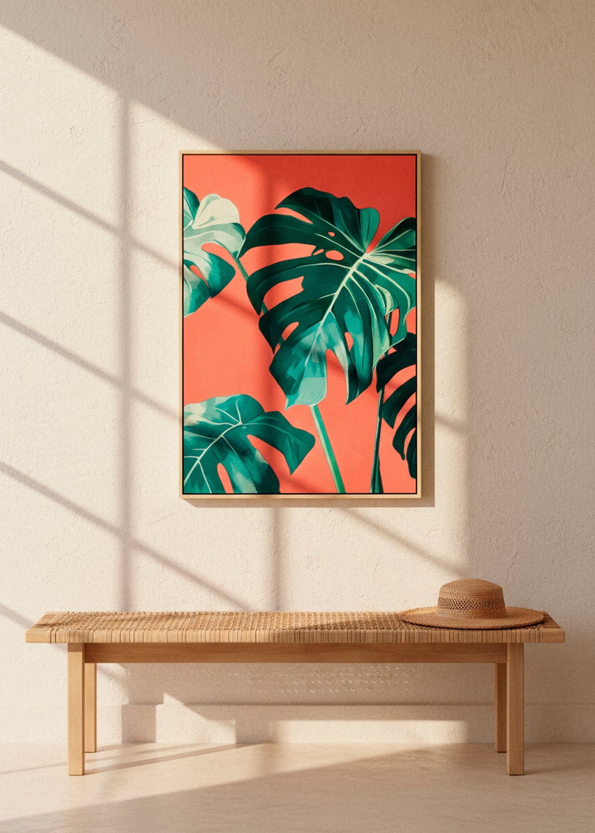 Framed “Monstera” canvas print with tropical leaves and bold coral tones, hanging above a woven bench in sunlit minimalist decor.