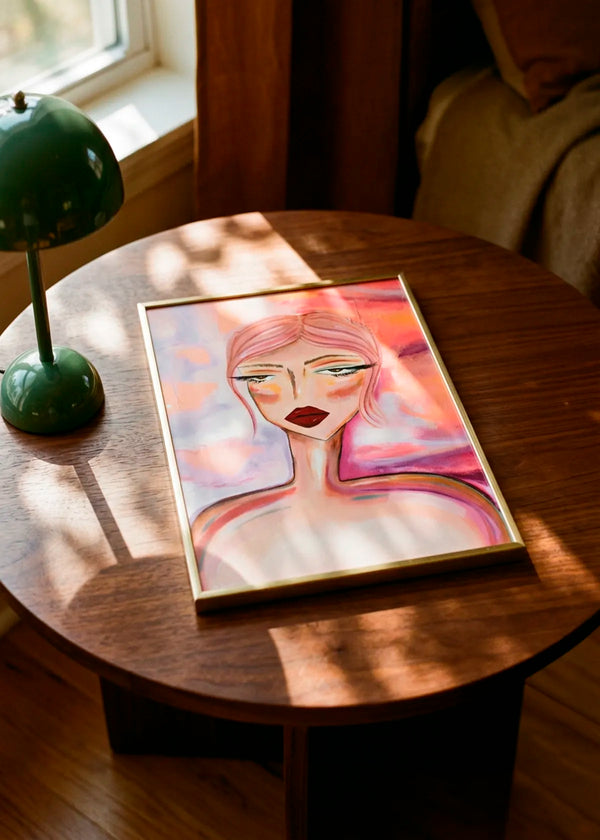 Moody Pink Hair Portrait by Amy Straley – framed expressive portrait poster featuring a woman with soft pink hair and deep red lips, styled on a round wooden table in a warm sunlit interior.