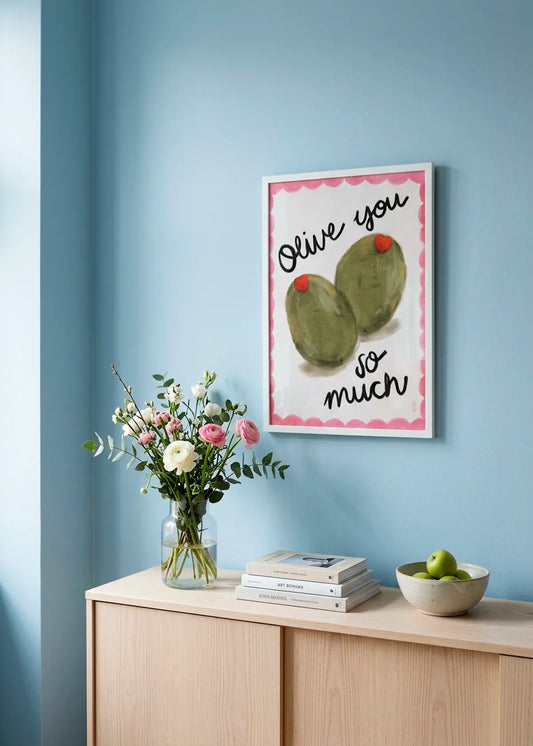 Olive You So Much by Dirkie Nell – framed playful olive poster with green olives, red hearts and pink border, styled on a soft blue wall above a light wood sideboard with flowers and books.