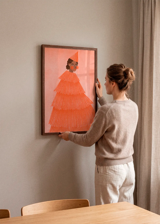 Orange Party Girl by Bea Müller art print featuring a woman in a vivid orange tiered dress and matching party hat against a soft pink background, displayed in a dark wood frame in a warm Scandinavian dining room.