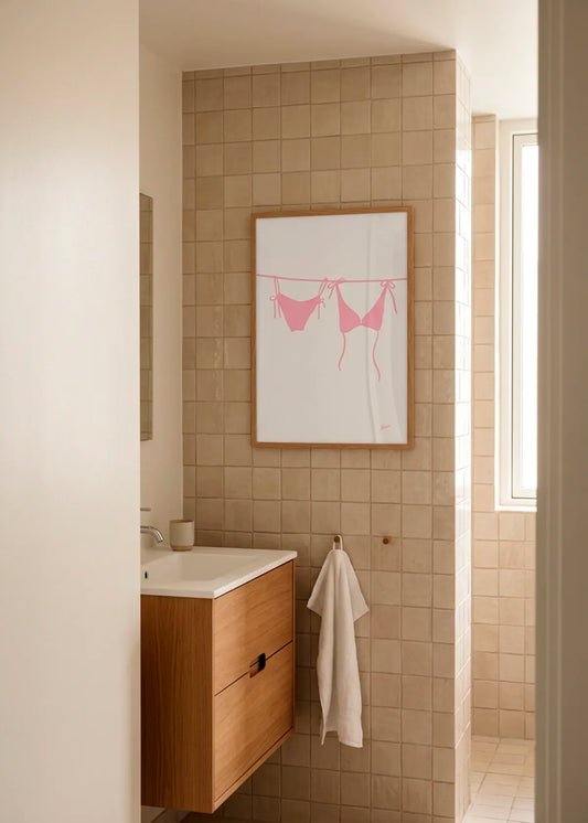 Pink Bikini by Athene Fritsch – framed minimalist art poster with a soft pink bikini illustration on an off-white background, styled in a warm neutral bathroom with beige tiles and natural wood.