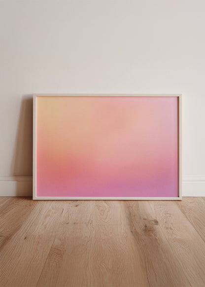 Framed abstract pink gradient poster with soft orange and violet tones, placed against a white wall on wooden flooring in a bright, modern room.