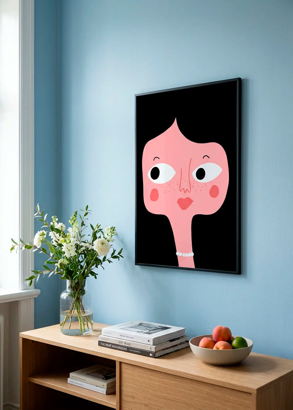 Quirky Pink Face by Athene Fritsch – framed illustrative portrait poster with a pink face on a black background, styled on a soft blue wall above a natural wood cabinet.