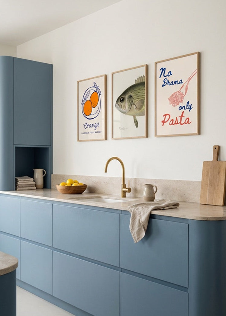 Three framed kitchen posters on a white wall above blue cabinets – oranges illustration, vintage fish print and “No Drama Only Pasta” typography in solid oak frames.