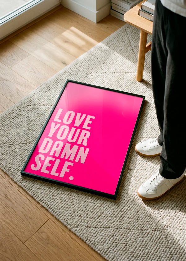 Self-Love Boldly by Athene Fritsch – framed hot pink typographic art poster with the words Love Your Damn Self, styled on a soft neutral rug in a bright modern interior.