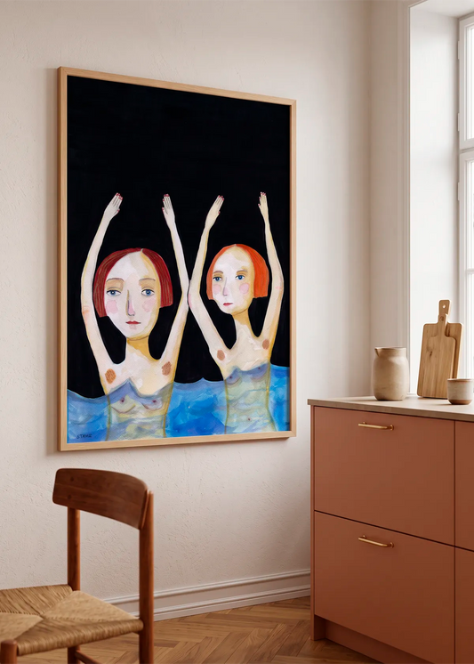 Synchronized Swimmers by Sharyn Bursic displayed in a natural wood frame on a soft cream wall beside warm terracotta kitchen cabinetry and wooden details.