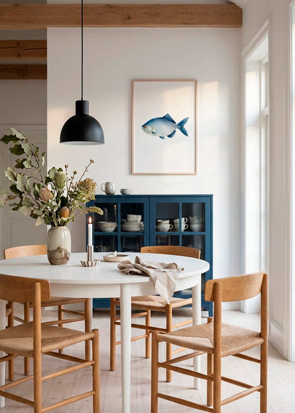 The Blue Fish art print in oak frame styled above a round dining table in modern Scandinavian dining room with blue cabinet and natural wood furniture