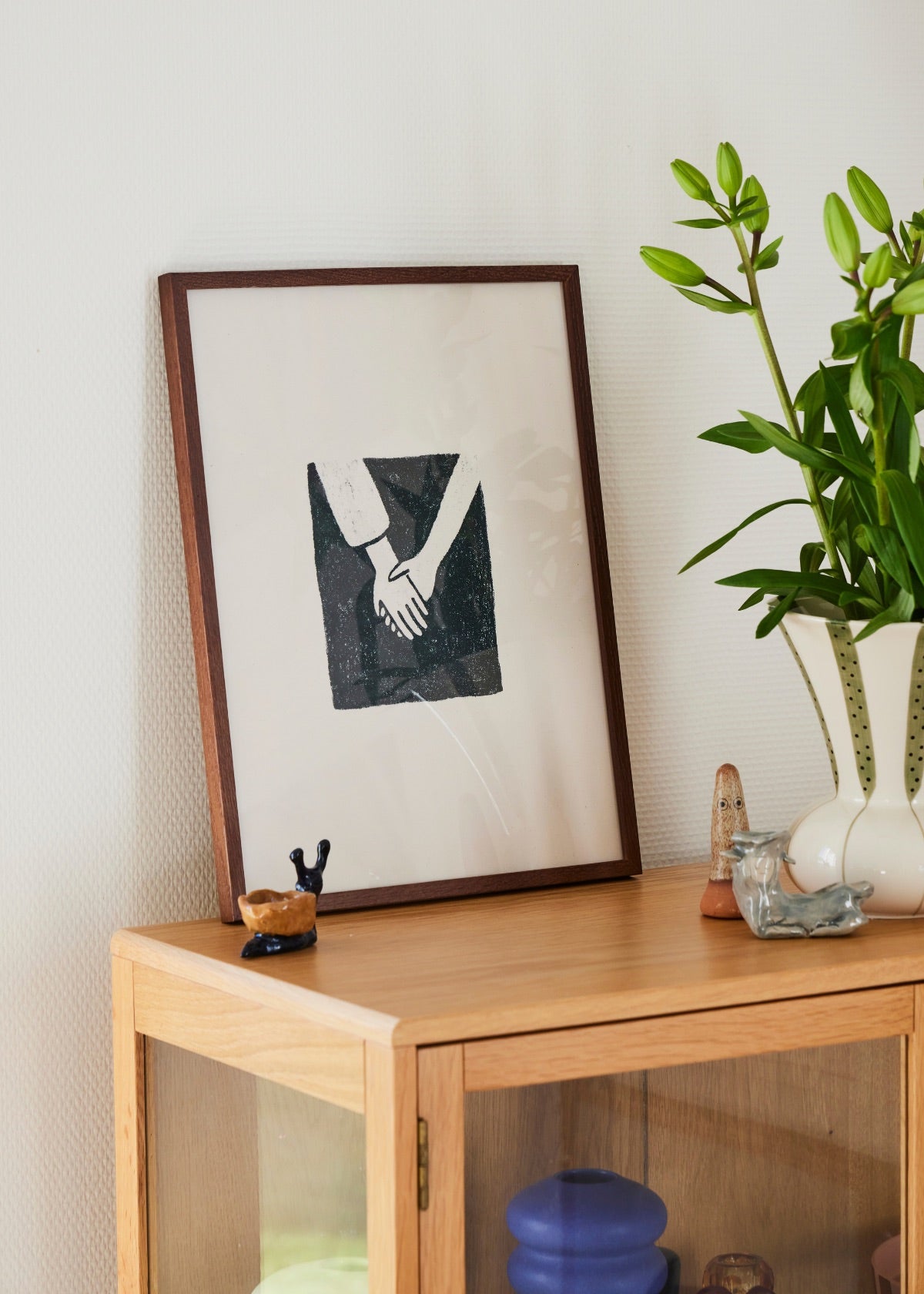 Framed minimalist poster of two hands holding, displayed on a wooden cabinet next to a vase with green flowers in a Scandinavian interior.