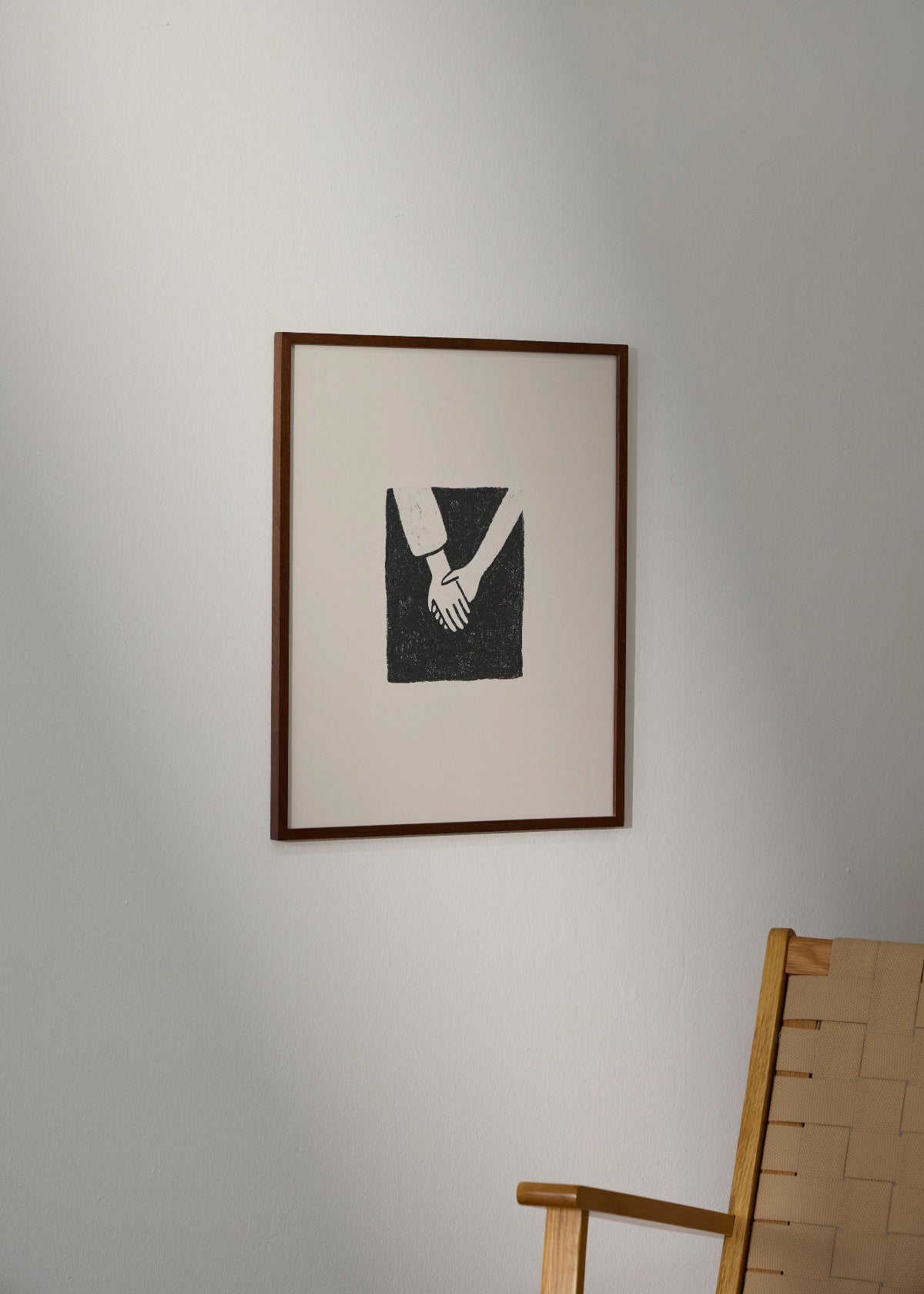 Togetherness poster from HelloPoster in dark wooden frame, minimalist black and white illustration of two hands holding, styled on a light wall with wooden chair.