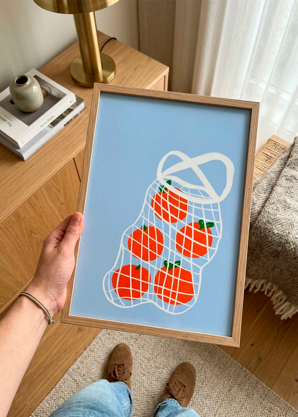 Tomatoes in Mesh by Athene Fritsch – framed illustrative art poster featuring red tomatoes in a white mesh bag on a soft blue background, held in a warm interior with natural wood details.