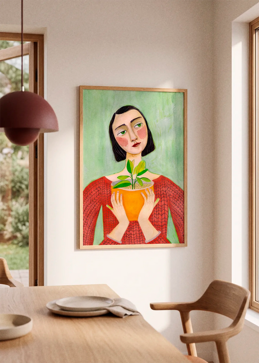 Woman Holding Green Plant by Sharyn Bursic displayed in a natural wood frame on a soft cream wall beside a light wooden dining table and chair in a bright, modern interior.