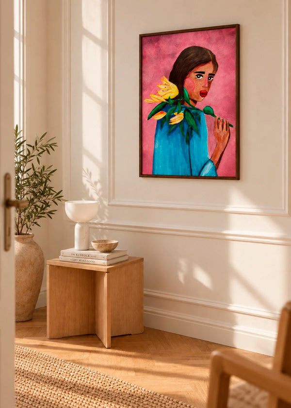 Woman With Lily plakat af Raissa Oltmanns i mørk træramme, stylet på en lys væg i et varmt interiør med træmøbler og naturligt sollys.