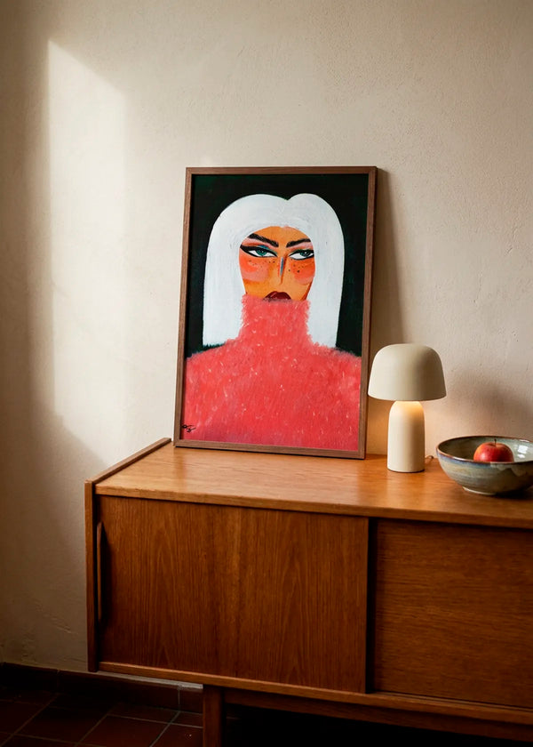 Woman in Pink Turtleneck by Amy Straley – framed portrait art poster featuring a woman with white hair and a textured pink turtleneck, styled on a warm wooden cabinet in a softly lit neutral interior.
