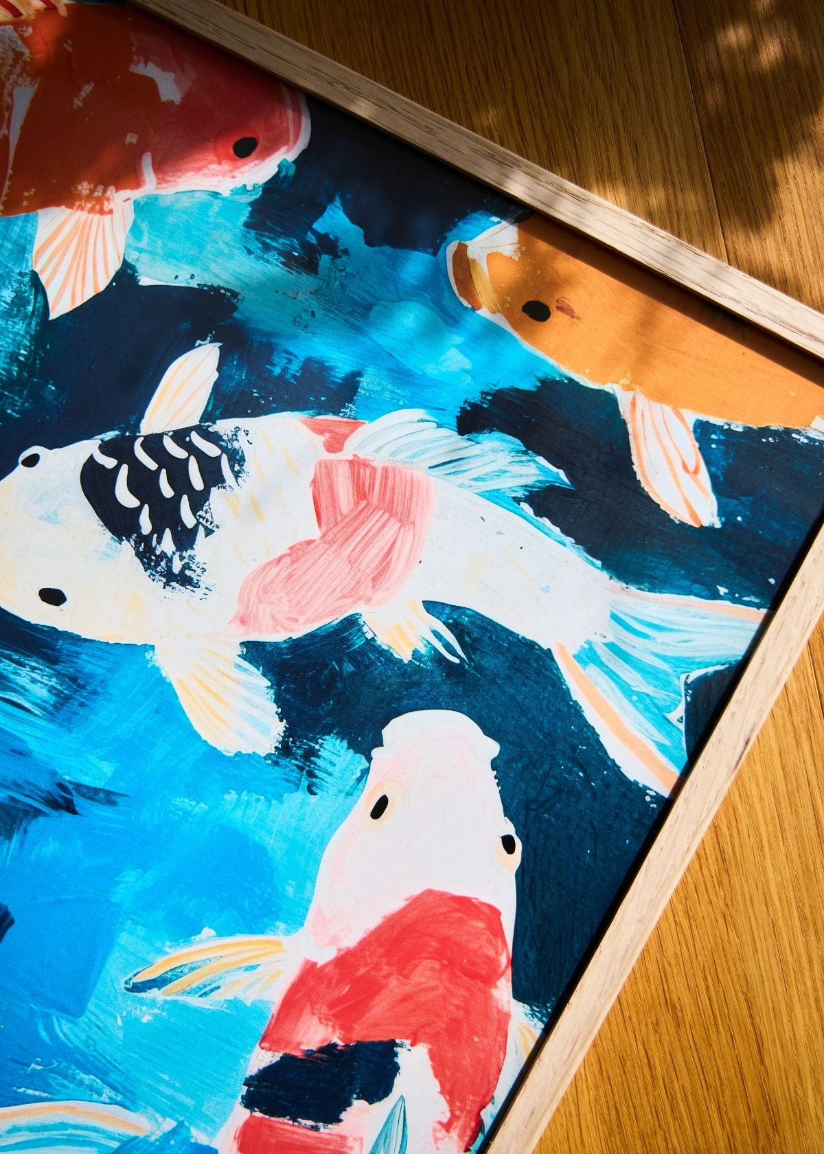 Framed “Koi” poster beside a ceramic koi sculpture on a wooden surface, with sunlight highlighting the bright aquatic colors.
